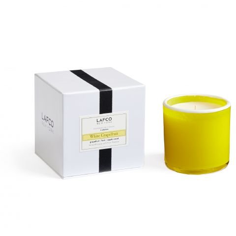 A yellow soy wax candle by Lafco in a glass holder sits next to a white box with a black stripe, labeled Lafco - White Grapefruit Classic 6.5 Candle. The scene is simple and minimalist, radiating subtle citrus charm.