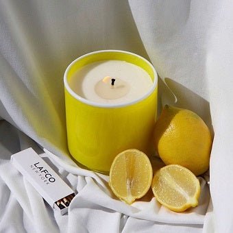 A Lafco - White Grapefruit Classic 6.5 Candle in a yellow holder rests on white cloth with whole and halved lemons, exuding citrus charm. A Lafco box nearby adds elegance to the fresh and bright scene.