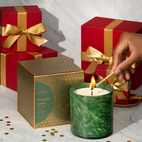 A hand lights the Lafco Woodland Spruce Signature 6.5oz Candle with a match as red gift boxes with gold ribbons and confetti dots sit in the background on a marble surface.