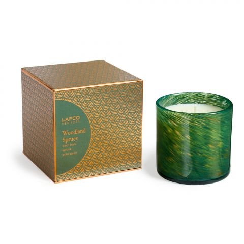 A green, textured Lafco Woodland Spruce Signature 6.5oz Candle in a glass holder sits next to a decorative green and gold box with geometric patterns and gold accents.