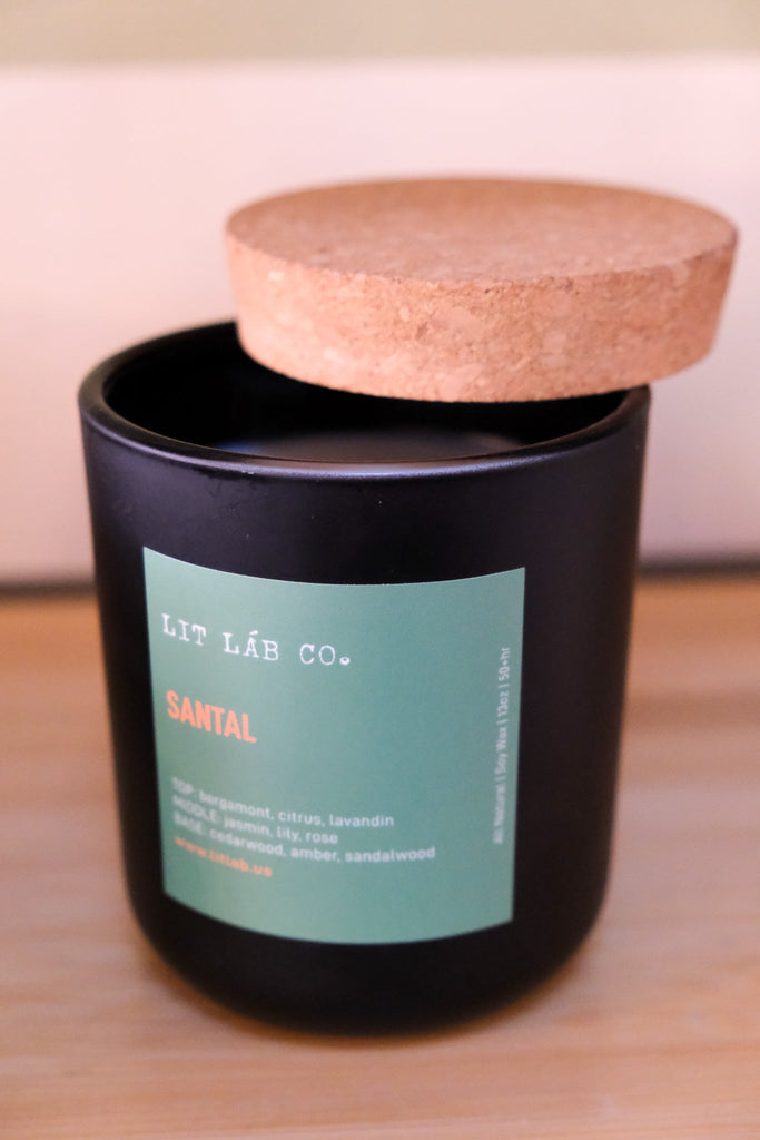 A black 13oz Santal candle from Lit Lab Co., with a wooden lid and wicks, sits on wood. Its label features scent notes of bergamot, citrus, lavender, rose, amber, and sandalwood. Crafted from soy wax for a clean burn.