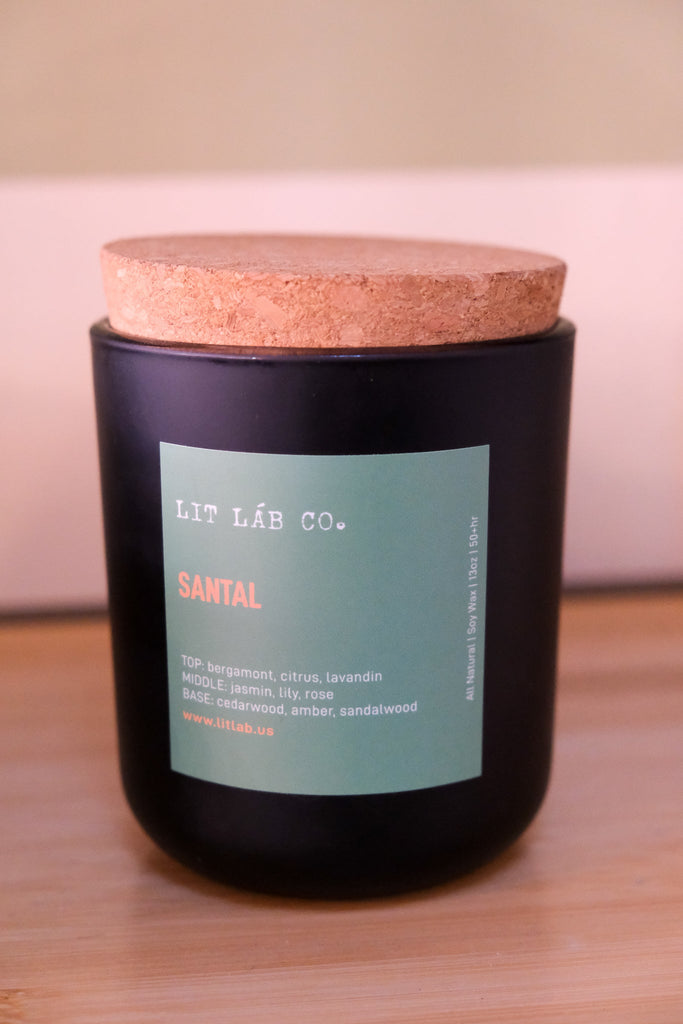 The Lit Lab Co. Santal 13oz candle features a black jar with a cork lid and wooden wicks for a clean burn. Its crafted from soy wax with fragrance notes: top (bergamot, citrus, lavandin), middle (jasmine, lily, rose), base (cedarwood, amber, sandalwood). Explore more at www.litlab.us.