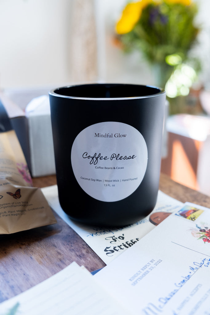 The Mindful Glow - Coffee Please Candle 13oz by Mindful Glow, a black jar candle made of coconut soy wax with a wood wick, sits on a table radiating a coffee aroma, surrounded by floral cards and cup with blurred flowers adding color in the background.