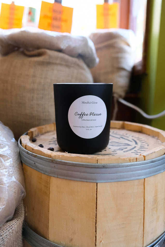 The Mindful Glow - Coffee Please Candle 13oz from Mindful Glow, crafted with coconut soy wax and a wood wick, rests on a wooden barrel surrounded by burlap sacks. Its black surface shines in sunlight streaming through the window.