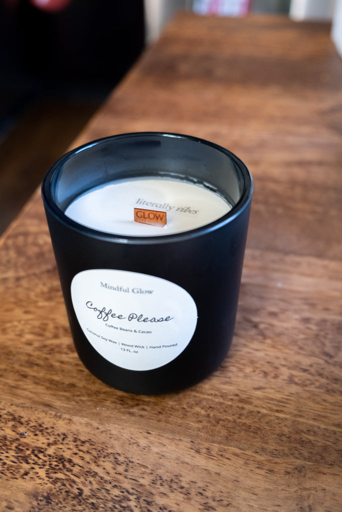 A Coffee Please candle by Mindful Glow, housed in a black jar on wood, reveals its wood wick and the phrase literally glowing engraved on a wooden piece inside. Made with coconut soy wax, it weighs 13oz.