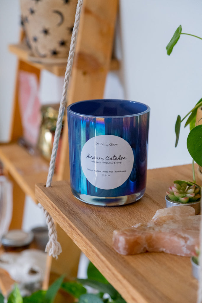 A blue coconut soy wax candle labeled Mindful Glow - Dream Catcher Candle 13oz by Mindful Glow rests in a glass jar on a wooden shelf. Around it are plants, a crystal, and decor items, with the shelf suspended by thick ropes.