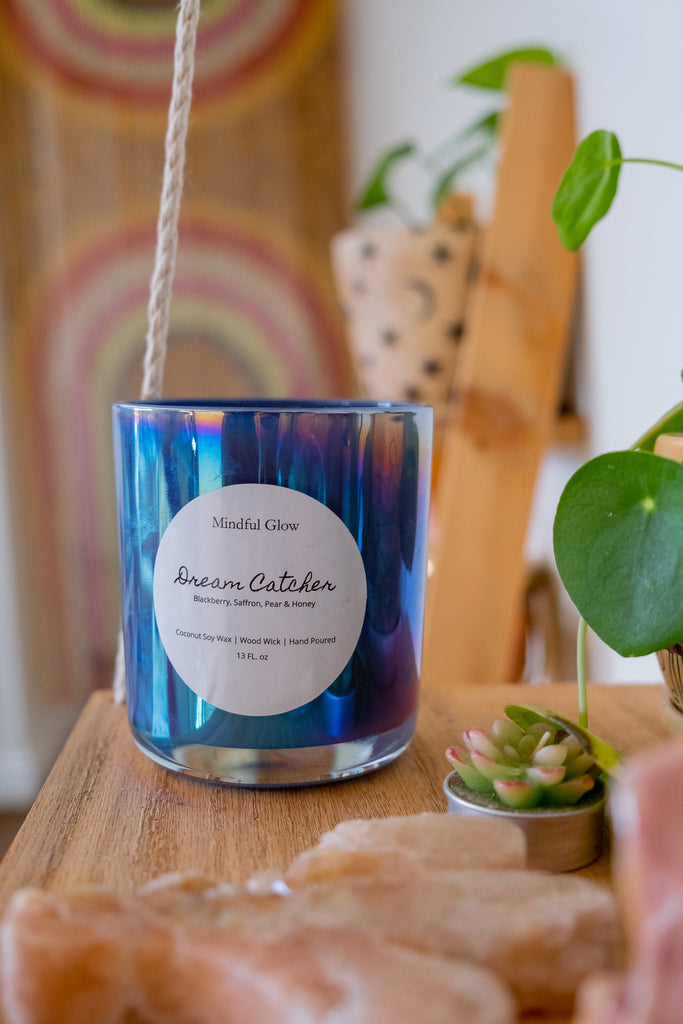 A blue and purple Mindful Glow - Dream Catcher Candle 13oz crafted from coconut soy wax rests on a wooden table, surrounded by small plants and crystals. The serene background features an abstract design and a potted plant.