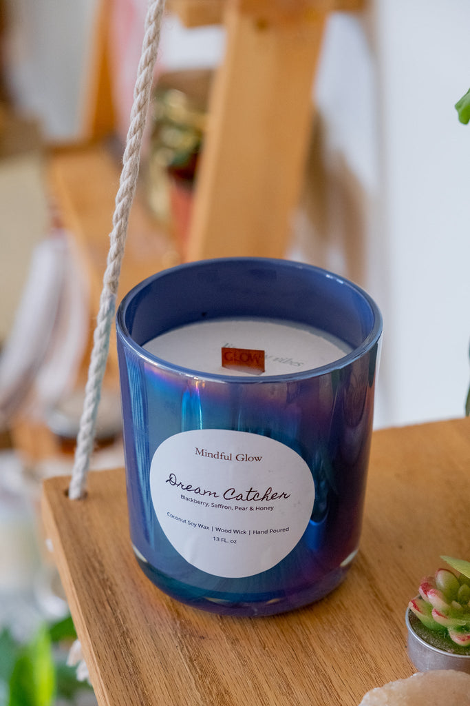 A 13oz glass candle labeled Mindful Glow - Dream Catcher sits on a wooden shelf, made with coconut soy wax and exuding rich scents of blackberry, jasmine, pear, and honey. Nearby, a small succulent plant peeks into view.