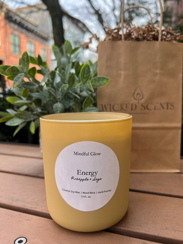 A 13oz yellow candle labeled Mindful Glow - Energy Candle crackles with a wooden wick on a table beside green leaves and a branded Wicked Scents paper bag. Made from coconut soy wax, the setting is outdoors among trees with a building in view.