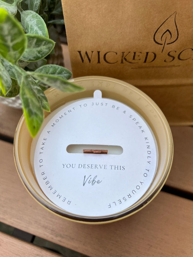 A round Mindful Glow - Energy Candle 13oz, featuring a wood wick and coconut soy wax, reads You Deserve This Vibe. Its lid says, Remember to take a moment to just be & speak kindly to yourself. A leafy plant and a paper bag with Wicked appear in the background.