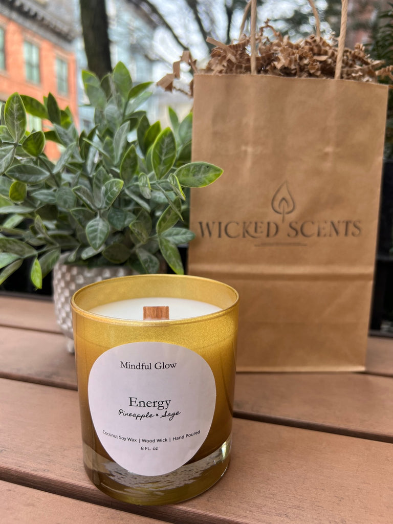 A Mindful Glow - Energy Candle 8oz from Mindful Glow, featuring coconut soy wax and a wood wick, emits pineapple and sage scents in a yellow glass jar. It rests on a wooden table beside a plant and WICKED SCENTS paper bag, with a window view enhancing the tranquil setting.