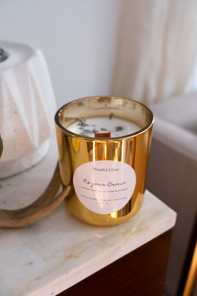 A soothing gold candle labeled Mindful Glow - Fix Your Crown Candle 13oz rests on a marble surface, hinting at its aromatherapy essence. Its accompanied by a modern white lamp and a glimpse of a beige armchair, crafting an oasis of calm elegance.