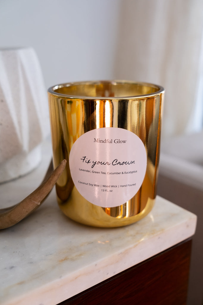 The 13oz Mindful Glow - Fix Your Crown candle, ideal for aromatherapy and relaxation, fills the air with lavender, green tea, cucumber, and eucalyptus. It sits elegantly on a marble-topped wooden surface beside a gracefully curved decor object.