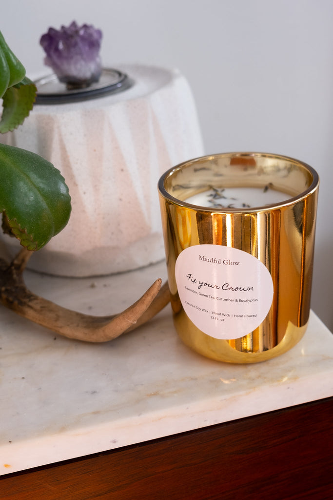 A gold 13oz Mindful Glow - Fix Your Crown candle rests on a marble surface beside a green plant and a stone piece with an antler, ideal for aromatherapy.