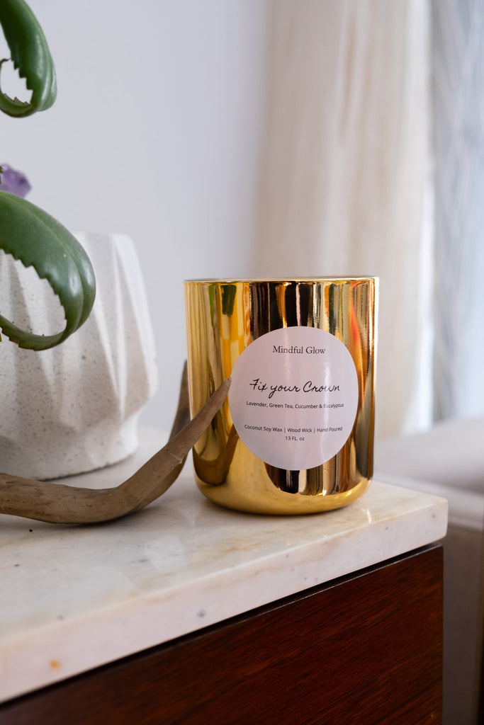 A 13oz gold Mindful Glow Fix Your Crown candle rests calmly on a marble-topped wooden surface, releasing a soothing scent. Nearby, a curved-leaf plant in a white geometric planter is framed by light curtains, enhancing the tranquil aromatherapy setting.