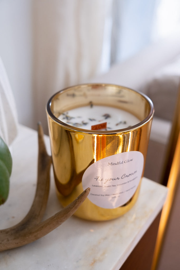 On a tabletop, the Mindful Glow - Fix Your Crown Candle 13oz in a gold holder relaxes with a white candle featuring a wooden wick and dried herbs. Nearby is a decorative antler, enhancing the aromatherapy ambiance.