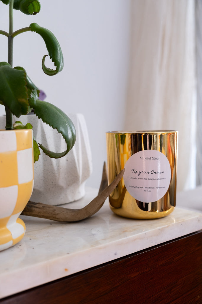 A gold Mindful Glow - Fix Your Crown Candle made from coconut soy wax rests on marble beside a checkered yellow pot holding a green plant. A decorative antler is close by, and a white curtain is partially visible in the background.