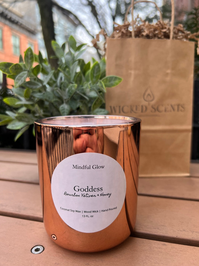 On a wooden table, a copper-hued Mindful Glow - Goddess Candle 13oz emits premium scents of Bourbon Vetiver + Honey. Accompanied by a Wicked Scents bag and lush plants, this coconut soy wax candle with wood wick creates an elegant backdrop.
.