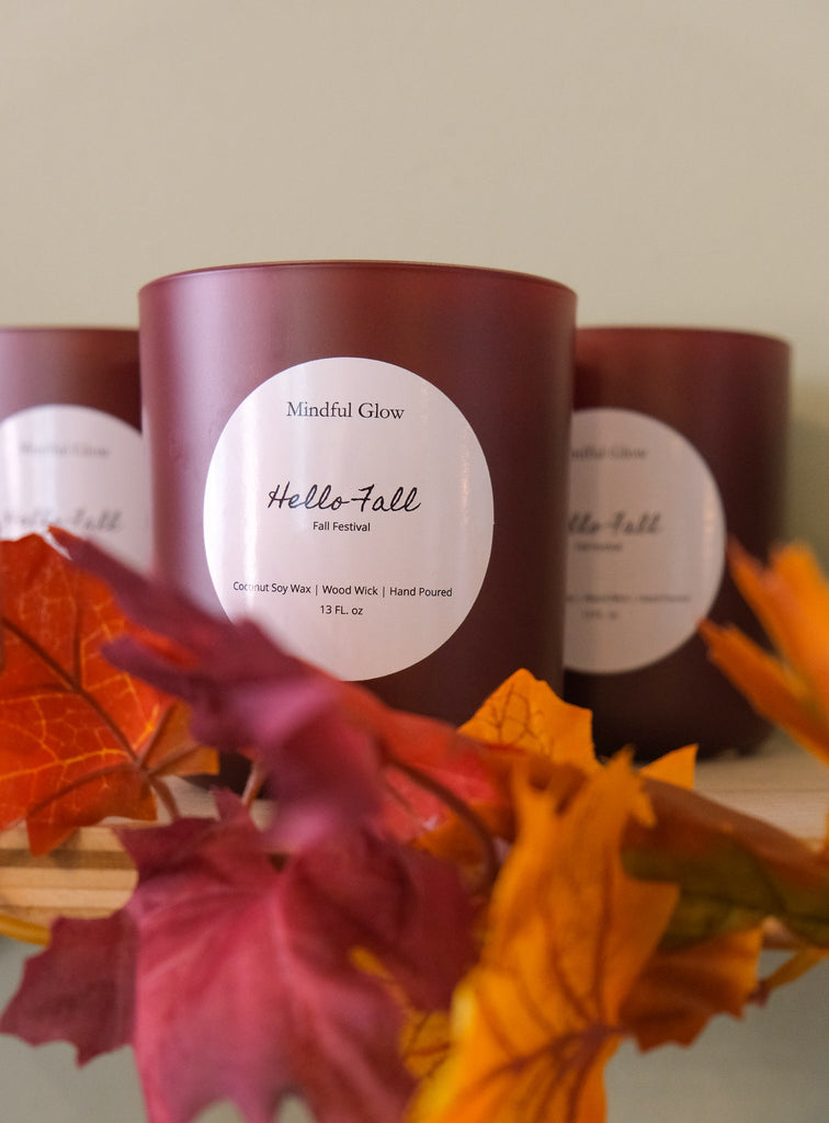 A brown 13-fl oz Mindful Glow - Hello Fall Candle made of coconut soy wax with a wood wick and blending cinnamon and vanilla sits on a shelf with colorful autumn leaves, surrounded by other enticing candles.