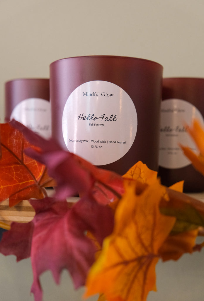 A burgundy Mindful Glow - Hello Fall 13oz candle, with cinnamon scent, is wrapped in artificial autumn leaves of orange, red, and yellow. Two similar citrus-scented candles are partially visible in the background.