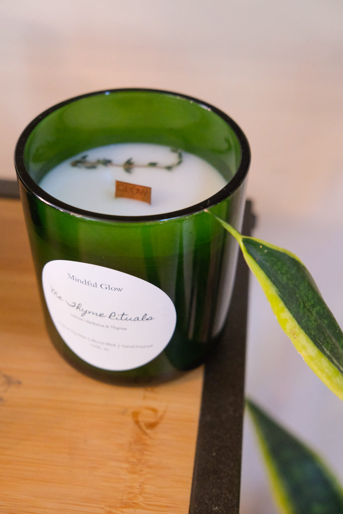 A Mindful Glow - Me Thyme Rituals Candle, crafted with coconut soy wax for a cleaner burn and fragranced with lavender, thyme, and citrus, sits on wood. The label marks Mindful Glow, while a plant with green and yellow leaves is partially visible next to the candle.