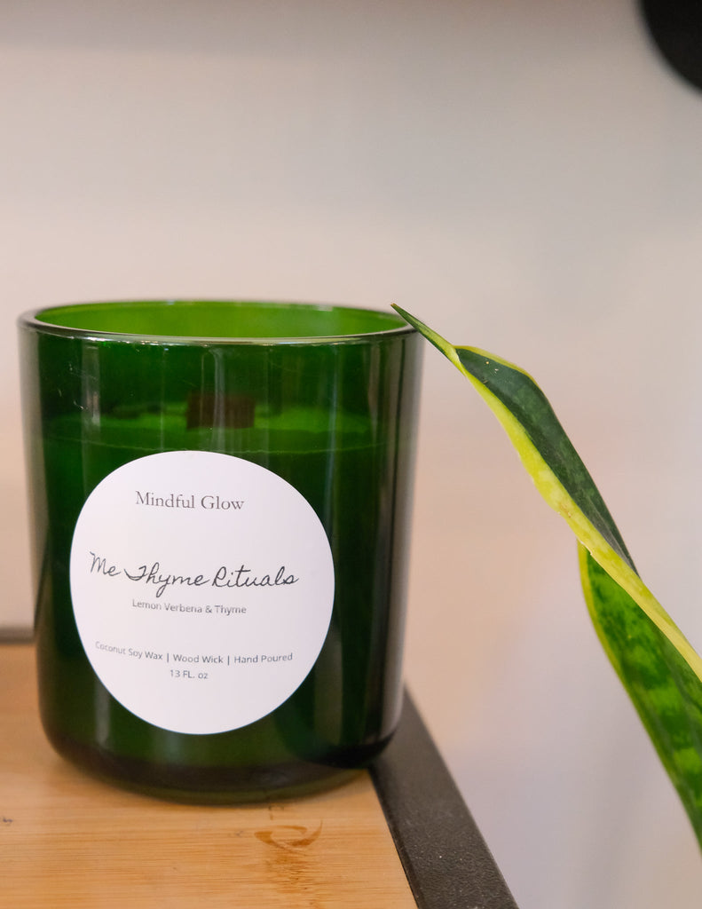 A green glass Mindful Glow - Me Thyme Rituals Candle 13oz by Mindful Glow, with premium Lemon Verbena & Thyme scents, coconut soy wax, and a wood wick, sits on a wooden surface. A leaf rests nearby.