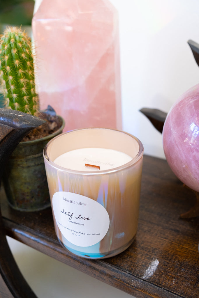 A lit Mindful Glow - Self Love Candle 13oz, crafted with coconut soy wax and a wooden wick, rests on a wooden table next to a small potted cactus and a large pink crystal, enhancing the tranquil atmosphere.