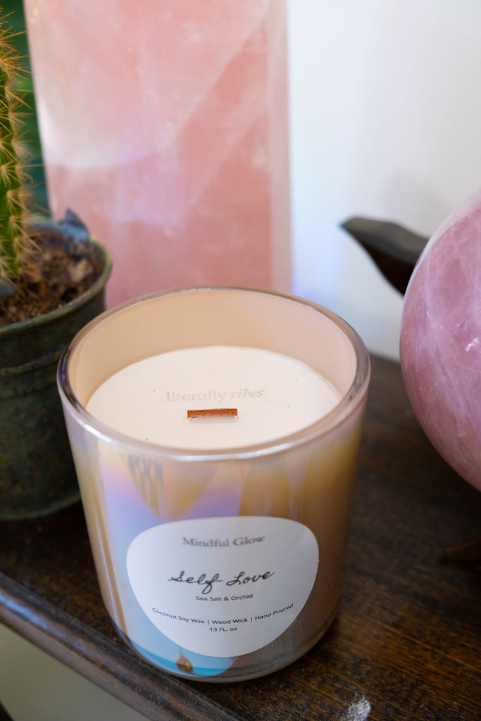 The Mindful Glow - Self Love Candle 13oz rests on a wooden surface near a potted cactus and a large pink crystal. This soy coconut wax candle with Salt & Orchid scent and a wooden wick is slightly burned, releasing fragrant sea salt and orchid notes.