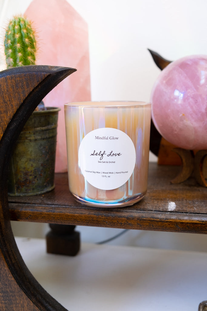 The 13oz Mindful Glow - Self Love Candle, made from coconut soy wax, rests on a wooden crescent-shaped shelf with a pink crystal and a cactus in a metal pot, creating a calming decorative vibe.