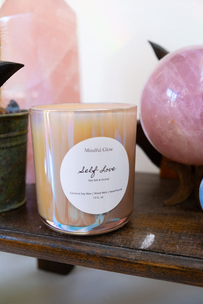 The Mindful Glow Self Love Candle, made from coconut soy wax, emits a calming sea salt and orchid scent. Its elegantly housed in a glass jar on a wooden shelf, complemented by a pink crystal sphere and potted plants.