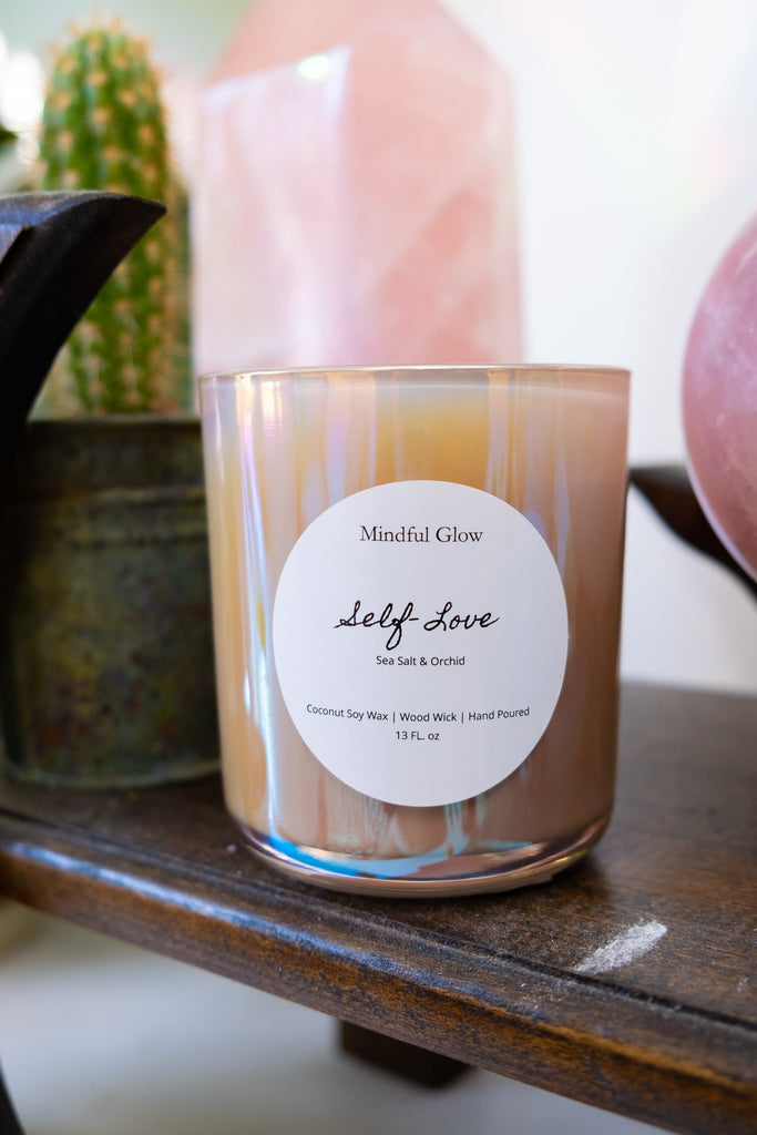 A Mindful Glow - Self Love Candle 13oz, made of coconut soy wax with a sea salt and orchid scent, rests on a wooden shelf. It is housed in a light iridescent container, decorated with cactus and pink crystals.