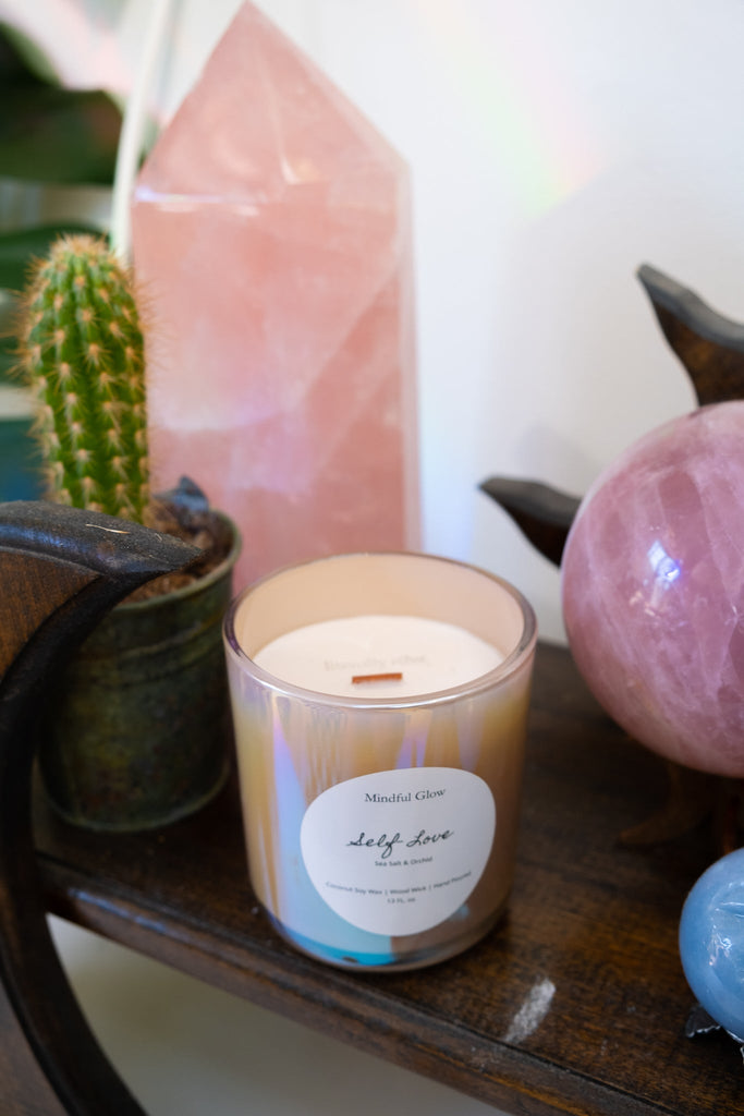 On a wooden shelf sits the Mindful Glow - Self Love Candle 13oz with a wood wick, next to a small potted cactus, large pink crystal, and pink sphere. Made from coconut soy wax, it enhances the tranquil and earthy ambiance with natural elements.