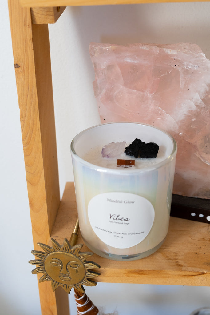 The Mindful Glow - Vibes Candle 13oz rests on a wooden shelf, crafted from coconut soy wax, beside a large piece of rose quartz. A brass sun decoration is partially visible underneath.