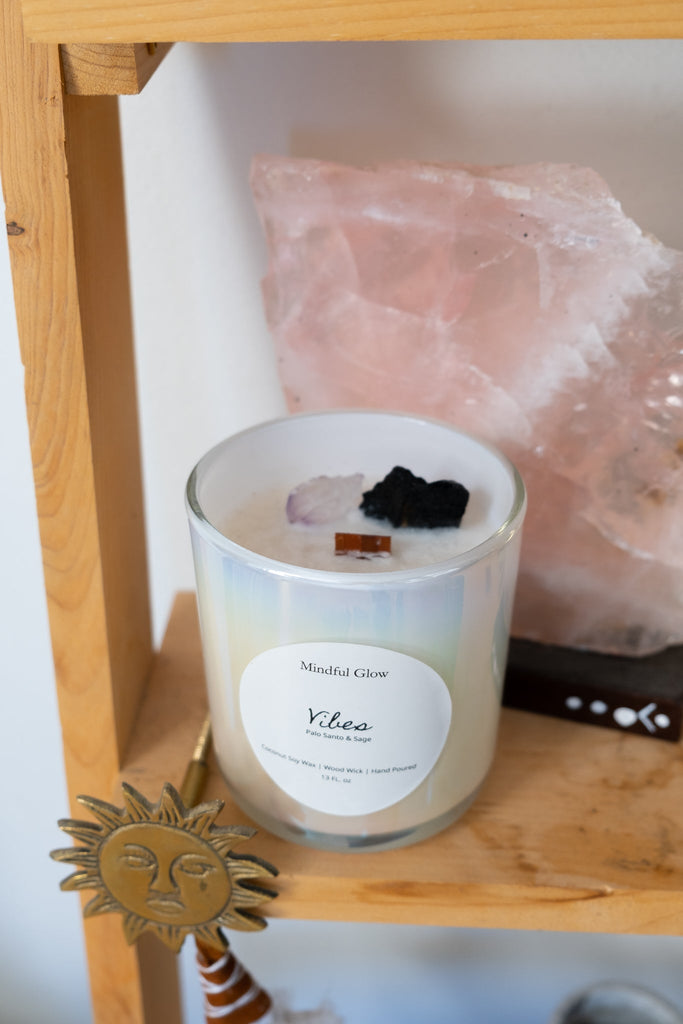 A 13oz Mindful Glow - Vibes Candle, crafted from soothing coconut soy wax, rests on a wooden shelf beside a large rose quartz emanating tranquility. A sun-shaped ornament at the edge of the shelf adds warmth and serenity to the scene.