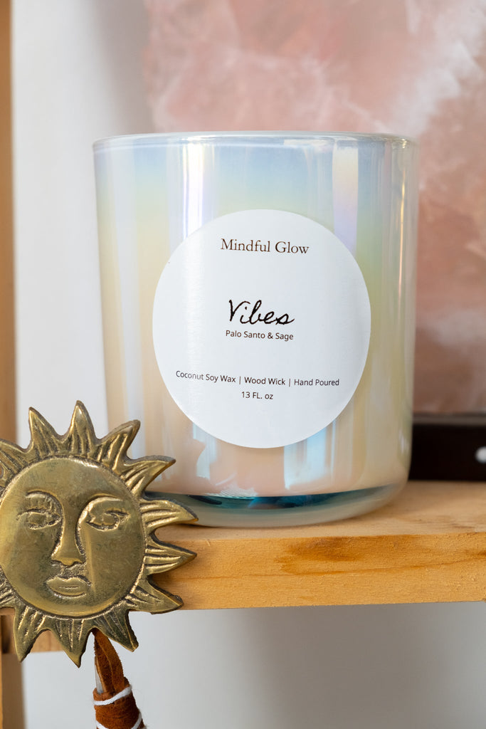 On a wooden shelf sits Mindful Glows Vibes Candle, 13oz, a white candle made from coconut soy wax with a wood wick. It exudes tranquility, while a partially visible decorative sun ornament on the left enhances the harmonious vibe.