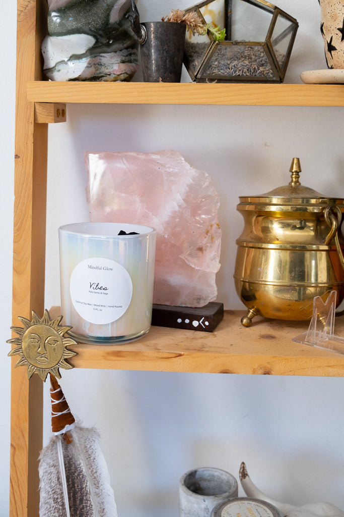 A wooden shelf displays treasures, including a Mindful Glow - Vibes Candle 13oz in a glass jar labeled Wildrose, made from coconut soy wax, a large rose quartz piece, a brass teapot, decorative feathers, and a front-attached sun ornament.