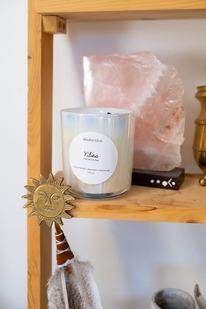 A wooden shelf displays a Mindful Glow - Vibes Candle 13oz made of coconut soy wax, next to a large pink crystal. Below the candle is a sun ornament, and partially visible in the background is a brown decorative item with painted dots.