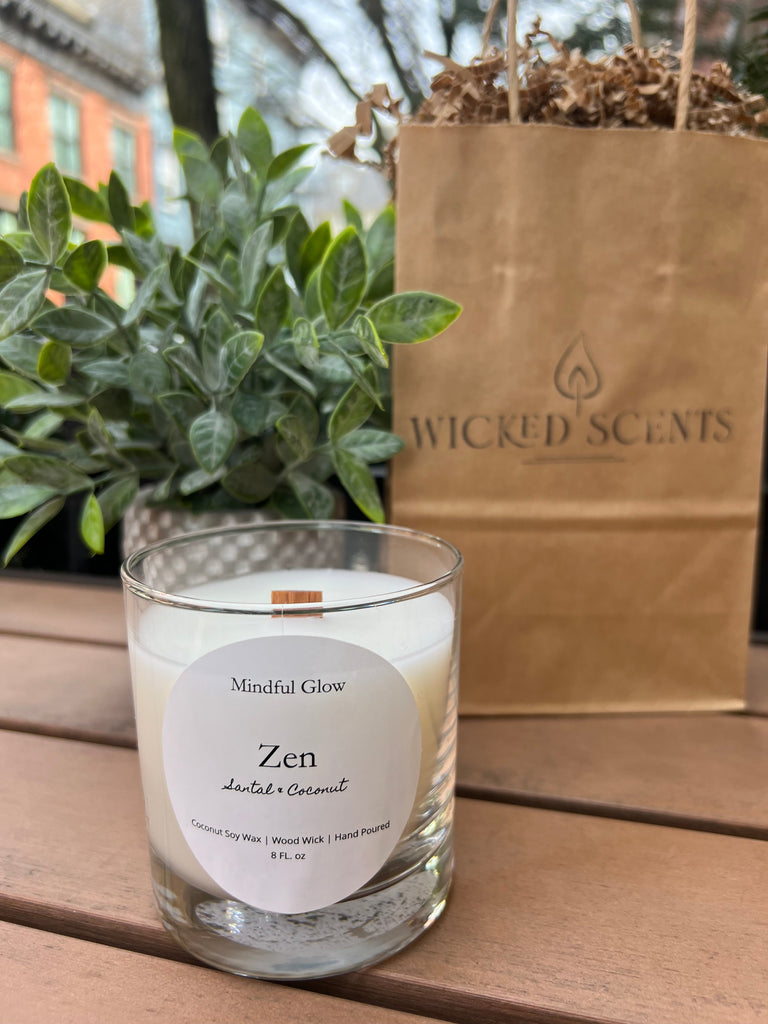 The Mindful Glow - Zen Woodwick Candle 8oz, a white coconut soy candle with a wooden wick, rests on a wooden surface beside green plants. Behind it is a brown Mindful Glow bag and an outdoor view of buildings and trees.