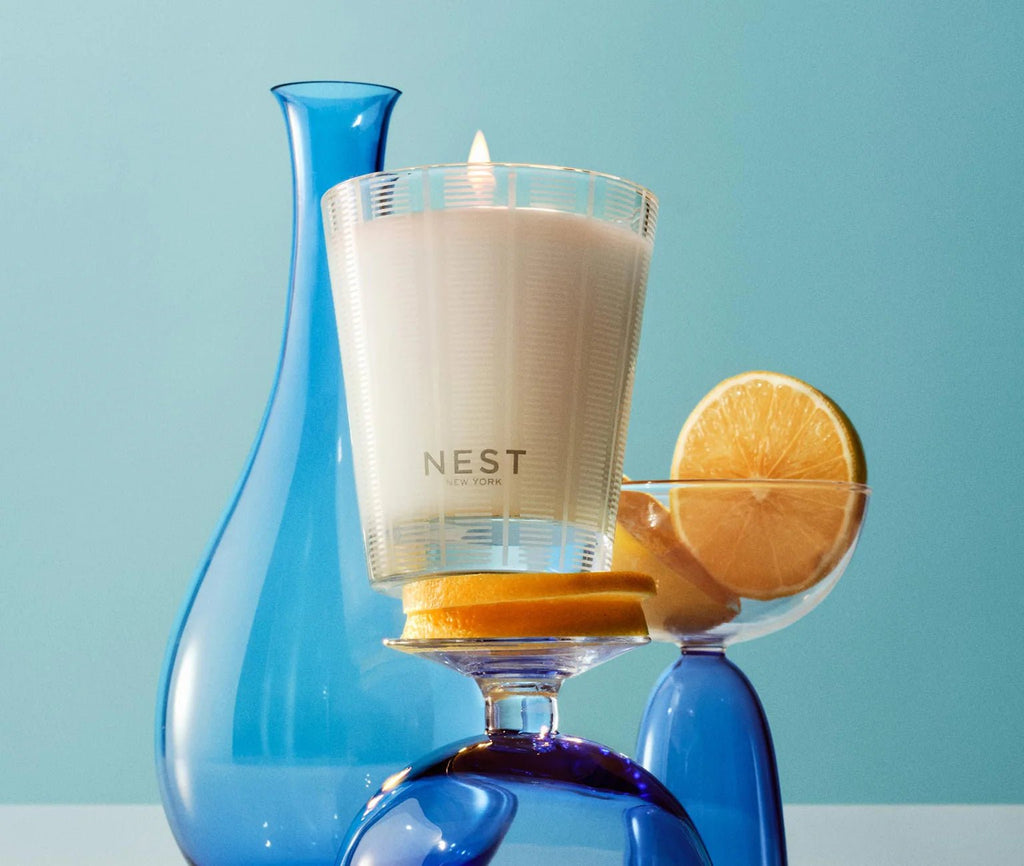 The Nest - Amalfi Lemon & Mint Classic Candle sits on an upside-down glass next to a sliced lemon in a cocktail glass, with a blue vase and bottle, all set against a light blue backdrop.