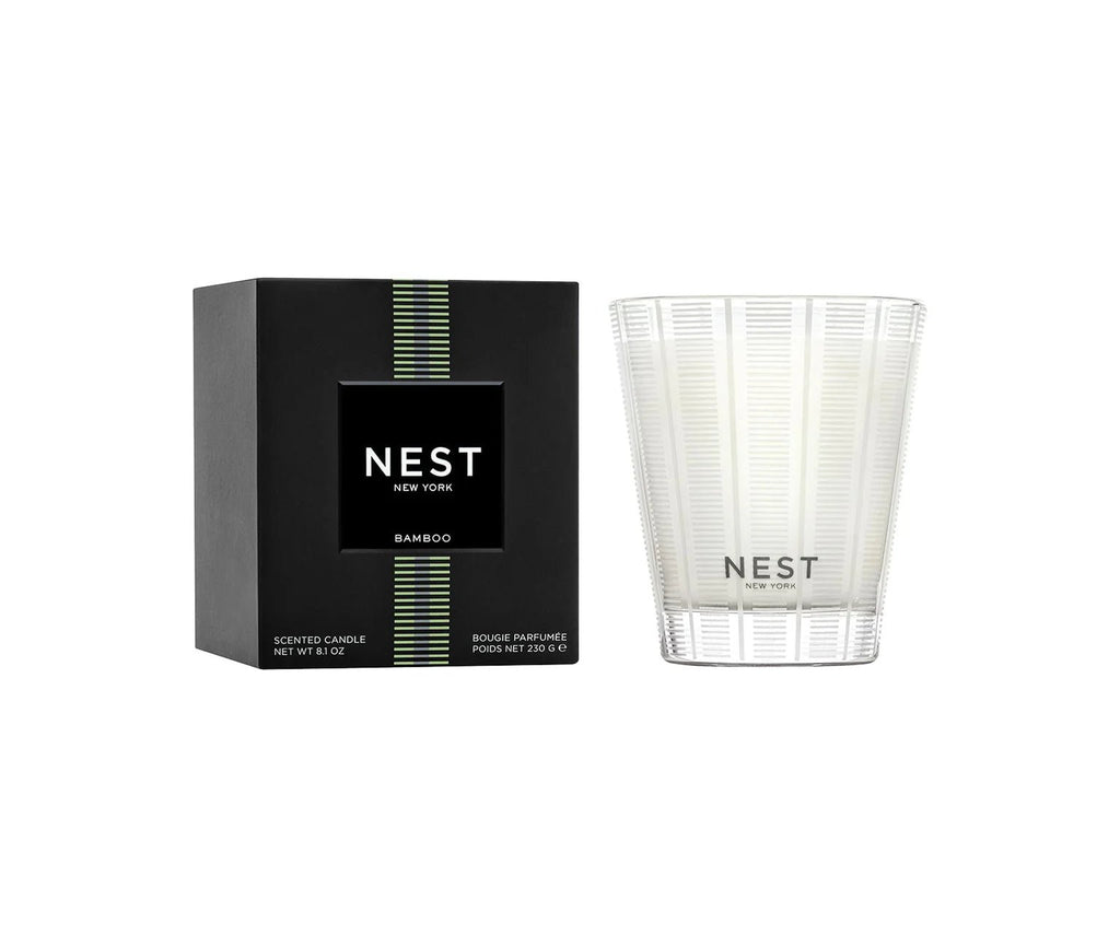 The Nest - Bamboo Classic Candle is displayed in a clear patterned glass jar beside its black box with green and white accents. This vegan, cruelty-free floral candle features the Nest logo and product details on both label and packaging.