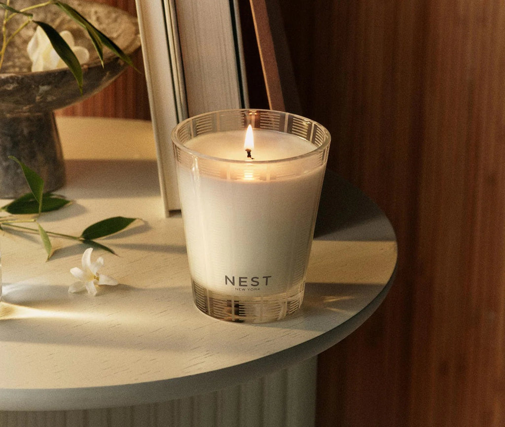 The Nest - Bamboo Classic Candle, a lit white floral candle in a clear glass holder labeled Nest, sits on a light round table with a flower, green leaves, and books in the background.