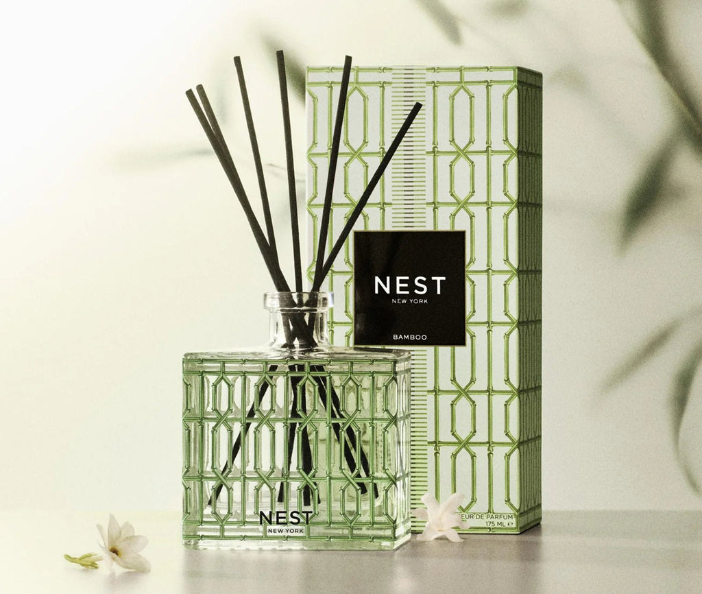 Nest - Bamboo Decorative Reed Diffuser features a green glass design and black reeds next to its patterned box. Softly blurred white flowers and green leaves in the background suggest a fresh floral fragrance.