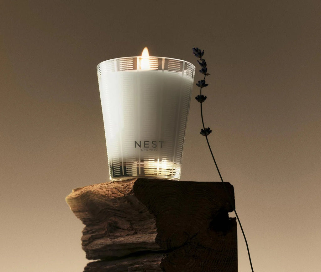 The Nest - Cedar Leaf & Lavender Classic Candle rests in a ribbed glass holder on rustic wood, its herbaceous scent with lavender notes filling the air as its flame casts a warm glow against a neutral backdrop.