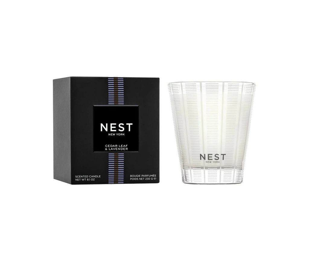 The Nest - Cedar Leaf & Lavender Classic Candle, in a clear patterned glass beside its black box with blue and silver stripes, is cruelty-free and fills your space with a refreshing herbaceous aroma.