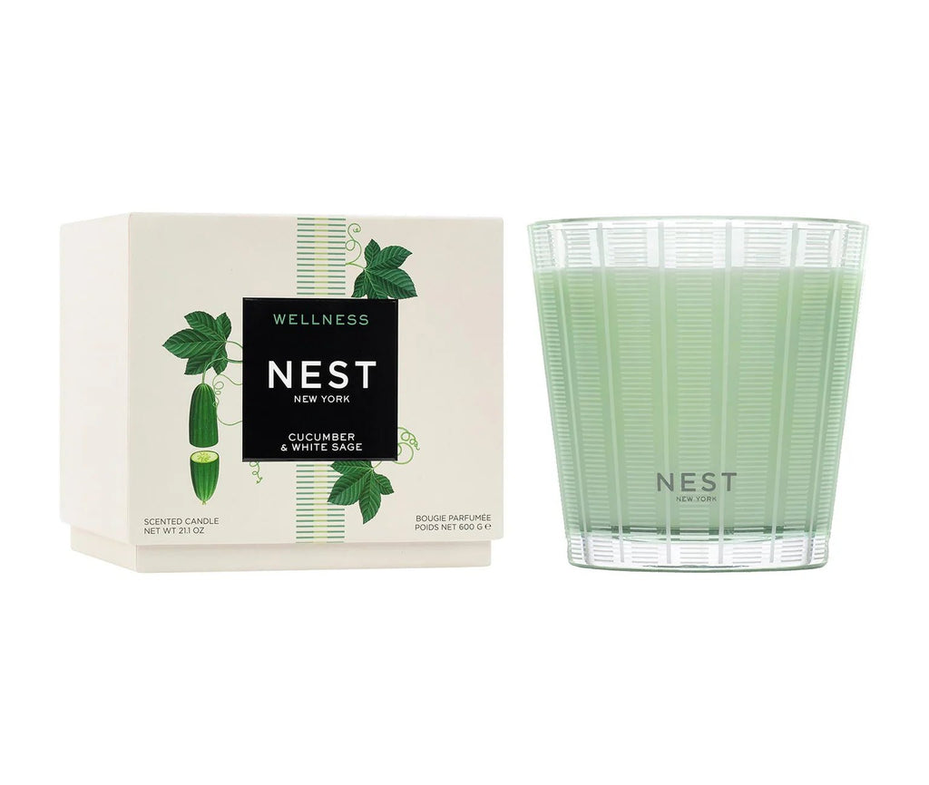 A Nest Cucumber & White Sage 3-Wick Candle in a clear glass holder sits beside its box, which displays cucumber and leaf designs.