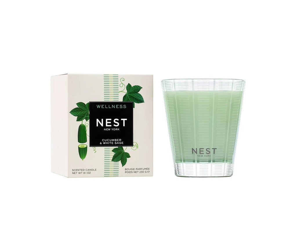 A light green Nest - Cucumber & White Sage Classic Candle sits in a clear glass holder beside its box, which displays cucumbers and green leaves to reflect the fresh, clean scent of cucumber and white sage.