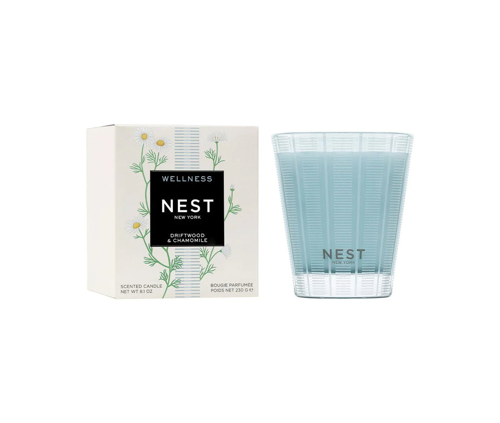 A light blue Nest - Driftwood & Chamomile Classic Candle in a textured glass holder sits next to its chamomile flower-decorated box, promising a clean burn and soothing scent.