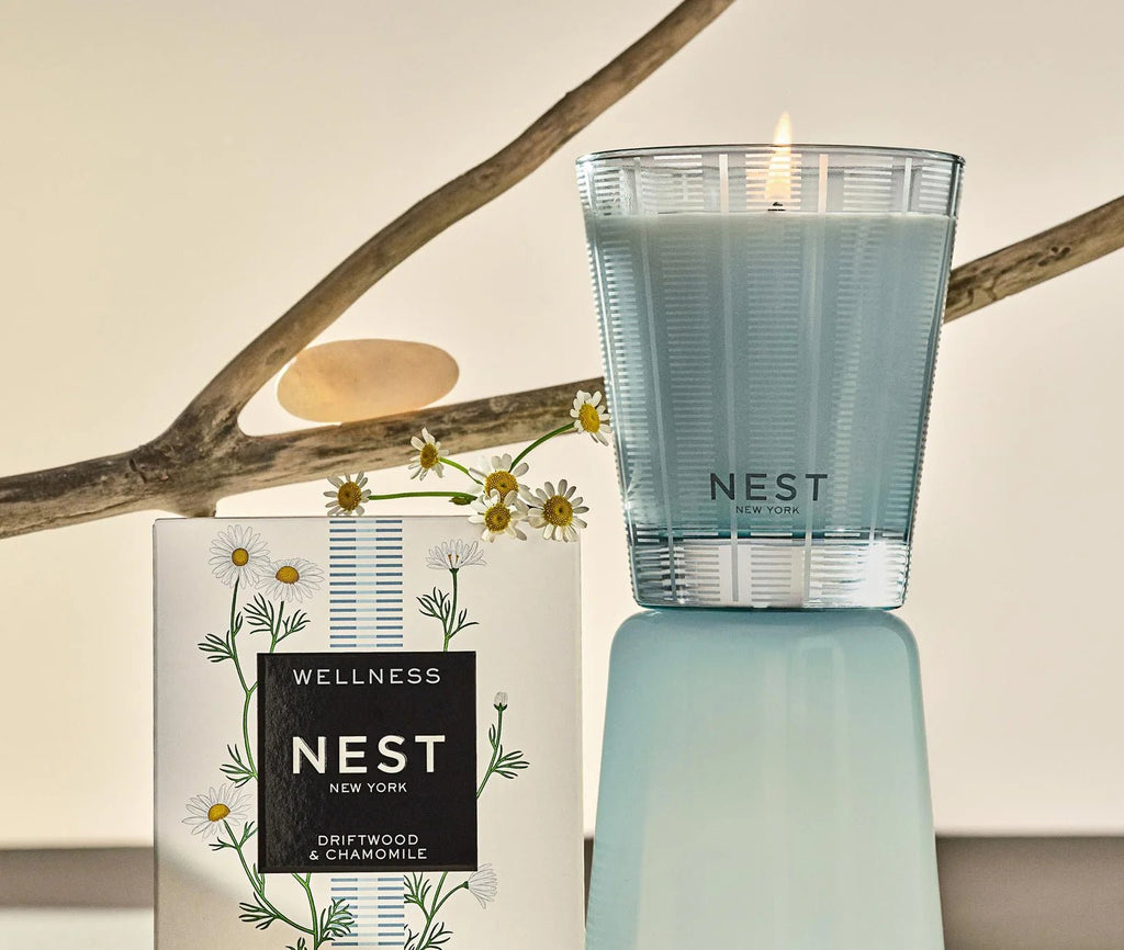 A lit Nest Driftwood & Chamomile Classic Candle sits on a pale blue pedestal next to its floral box, with a branch and chamomile flowers in the background, emphasizing its soothing scent and clean burn.