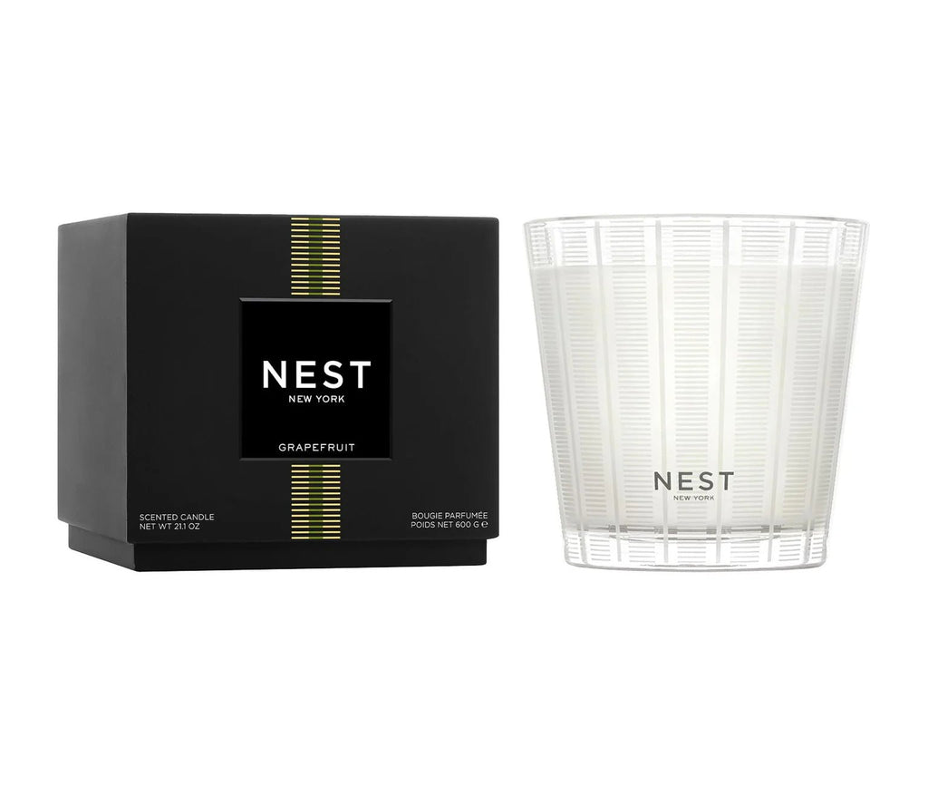 The Nest - Grapefruit 3-Wick Candle features a clean burn in a clear, ribbed glass jar and comes in sleek black packaging with the Nest logo and gold stripes—a luxurious fragrance addition to any space.