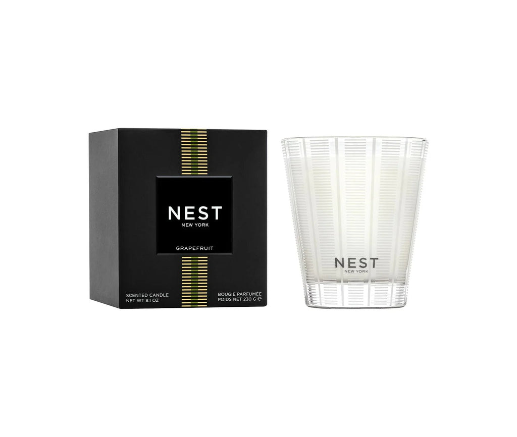 The Nest - Grapefruit Classic Candle by Nest features a pink grapefruit scent and comes in a clear patterned glass, displayed next to its black box adorned with gold stripes and white text.